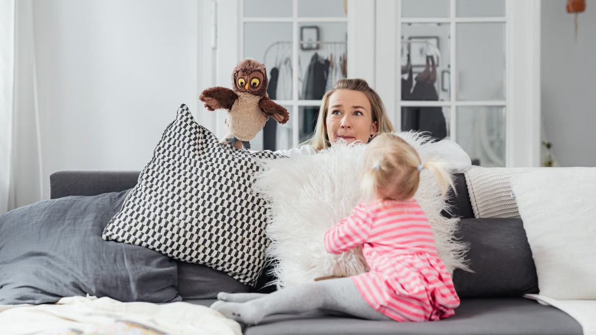 A woman and her daughter play with an owl puppet on a cozy couch in a bright living room.